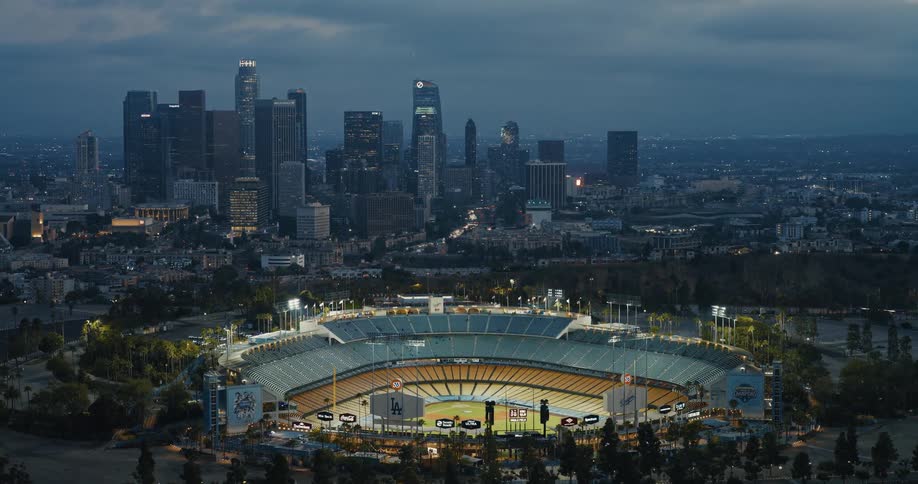 A slow-pulling aerial shot reveals the iconic Dodger Stadium illuminated at dusk with the Downtown Los Angeles skyline in the background