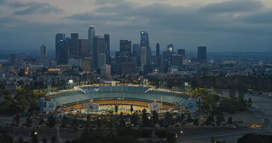 A slow aerial pull-back reveals the iconic Dodger Stadium illuminated at dusk with the Los Angeles city skyline in the background