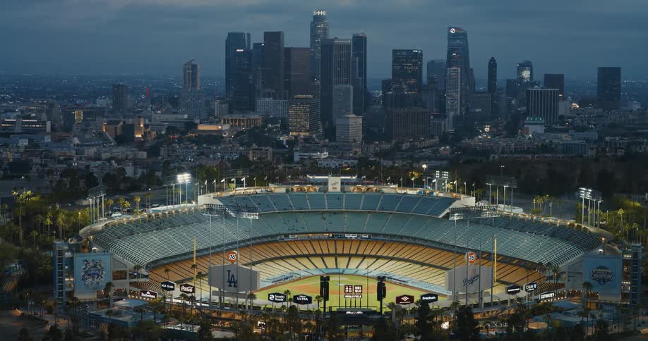 A stunning aerial view captures the illuminated Dodger Stadium at dusk with the sprawling Los Angeles city skyline in the background