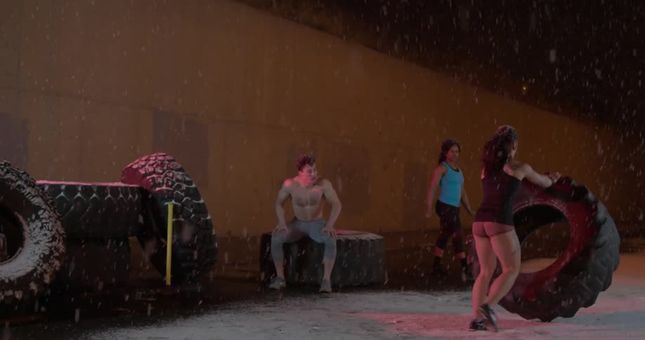 A group of people flip large tires outside while it snows