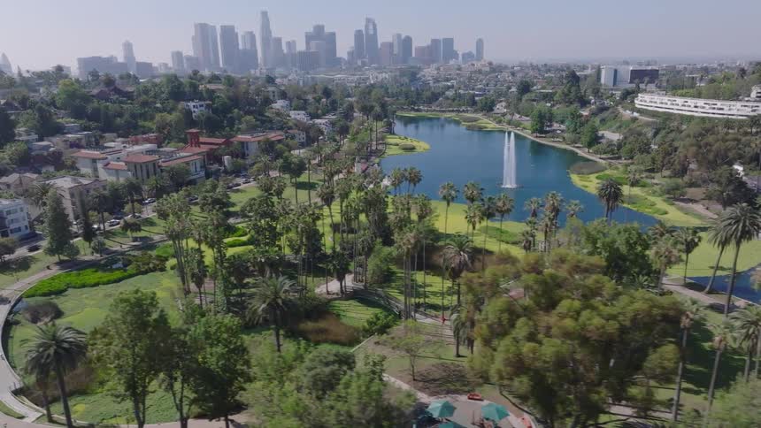 An aerial dolly shot flies over the lush green trees of Echo Park, showing the lake and fountain with the downtown Los Angeles skyline in the background
