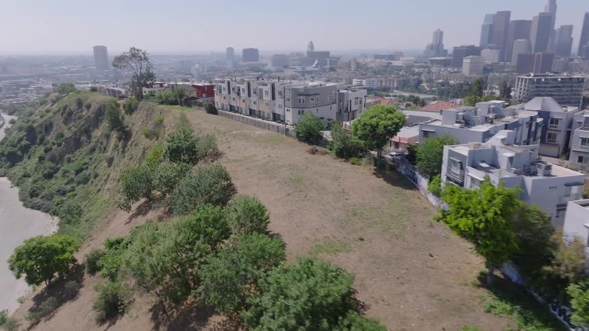 An aerial shot flies over a hillside residential area, revealing the downtown Los Angeles skyline on a clear, sunny day
