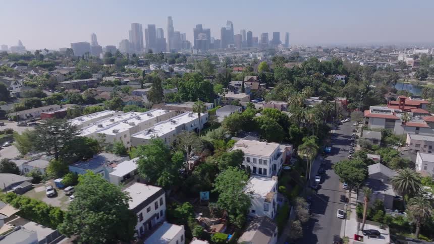 A slow aerial view drifts over a quiet residential neighborhood and park, revealing the vast Los Angeles city skyline in the background on a sunny day