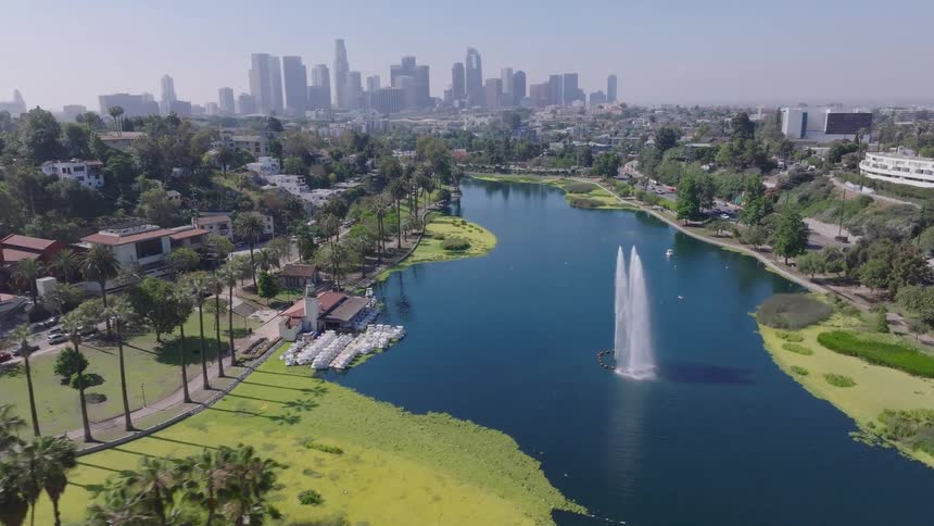 A scenic aerial flight over the lush palm trees and green landscape of Echo Park Lake towards the fountain, with the Downtown Los Angeles skyline in the distance