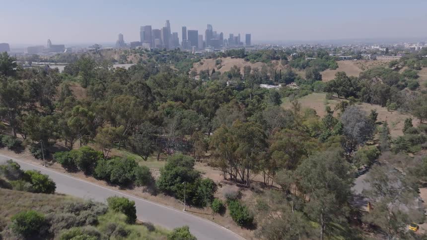 Aerial drone shot flies over a playground and lush green park towards the distant downtown Los Angeles skyline on a sunny day