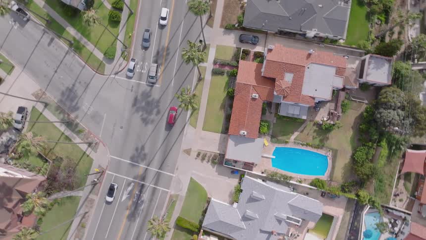 Top-down aerial footage slowly zooms in on a sun-drenched suburban intersection, revealing houses, swimming pools, and daily traffic