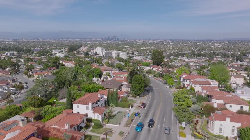 Aerial drone footage flies over a sunny suburban neighborhood street with cars, revealing the sprawling Los Angeles cityscape and downtown skyline in the distance