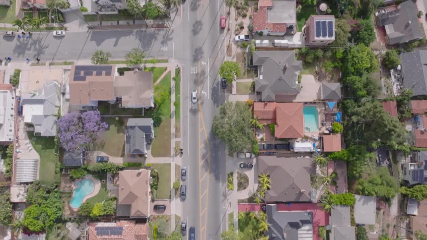 An aerial top-down drone shot follows a road through a lush, green suburban neighborhood with tile-roofed houses and swimming pools