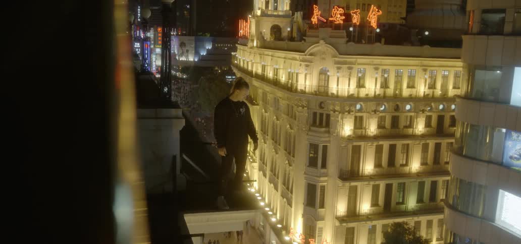A man balances on the ledge of a rooftop overlooking a city