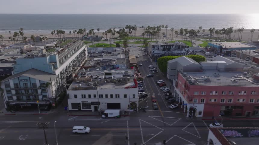 An aerial shot slowly pans left over the streets and buildings of Venice Beach, California, with the Pacific Ocean and palm trees in the background