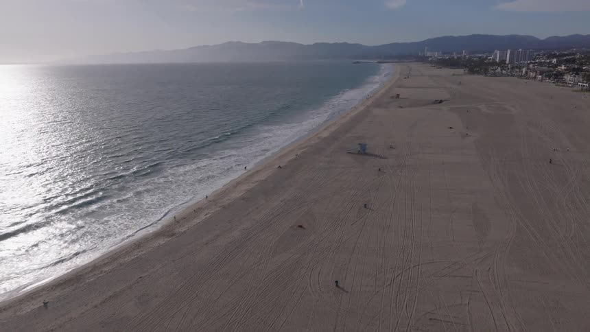 Aerial footage glides slowly over the vast, sandy shores of a California beach as gentle waves roll in from the ocean