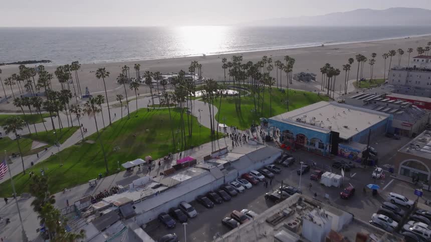 A stunning aerial view flies over the iconic Venice Beach, revealing the famous skate park and boardwalk with the sun reflecting on the Pacific Ocean