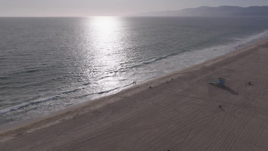 A slow-pulling-back aerial shot reveals a vast, serene beach with the sun reflecting on the ocean's surface