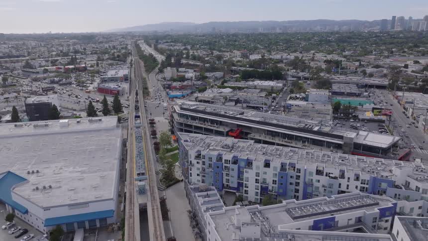 Aerial footage follows an elevated train moving through a modern urban landscape with a highway and city skyline in the distance