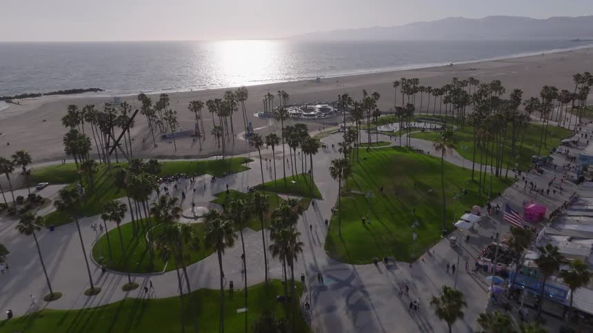 Sweeping aerial footage flies over the iconic Venice Beach boardwalk, park, and skatepark towards the Pacific Ocean at golden hour