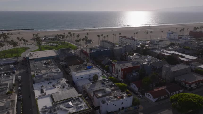 An aerial view of the cityscape, palm trees, and the sandy shore of Venice Beach, California, with the sun reflecting on the Pacific Ocean