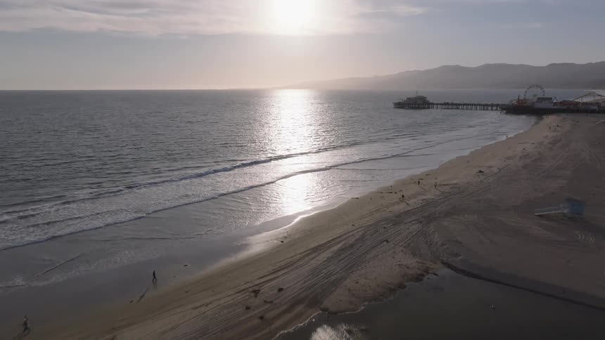 An aerial view flies over the sandy beach and out to the Pacific Ocean with the Santa Monica Pier visible in the distance at sunset