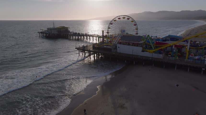A scenic aerial flight towards the iconic Santa Monica Pier at sunset, featuring the famous Ferris wheel and amusement park over the Pacific Ocean