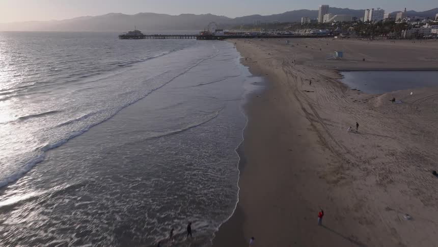 An aerial view captures gentle waves lapping the shore of Santa Monica beach with the iconic pier visible in the distance at dusk