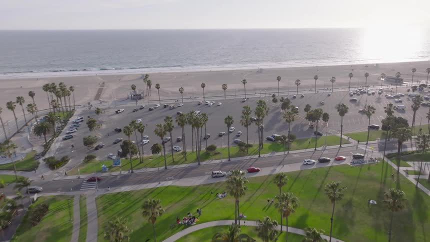 Aerial drone footage flies over lush green trees and a park towards the sunny Santa Monica Beach in California