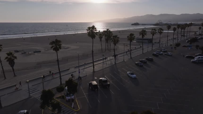 Soaring over the palm trees and parking lot towards Santa Monica Beach and the iconic pier as the sun sets over the Pacific Ocean