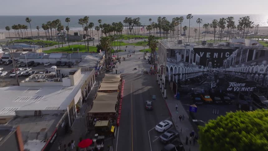 An aerial view flies down a main street towards the iconic Venice Beach boardwalk and the Pacific Ocean
