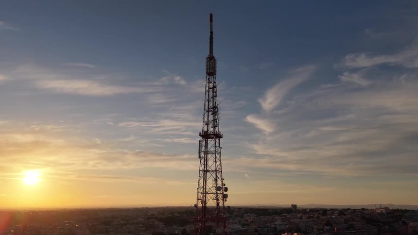 An aerial shot slowly pulls back from a tall telecommunications tower silhouetted against a stunning golden sunset over a city
