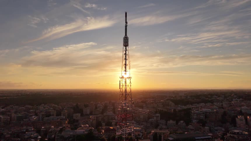 A dramatic aerial view shows a large telecommunications tower silhouetted against a beautiful golden sunset over a sprawling city