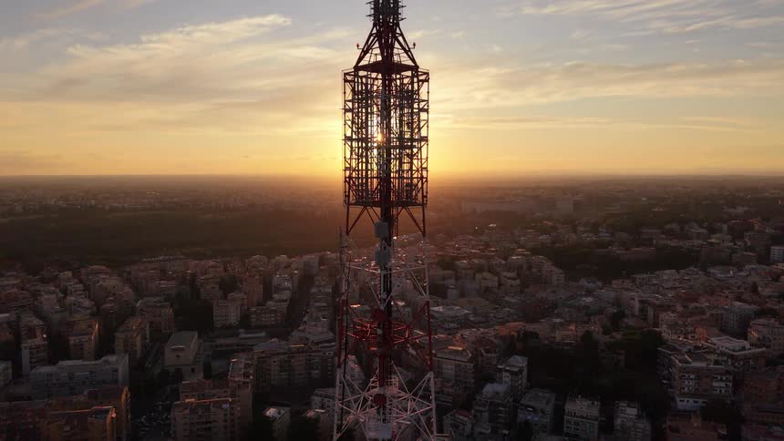 An aerial view ascends a massive telecommunications tower silhouetted against a beautiful sunset over a vast cityscape
