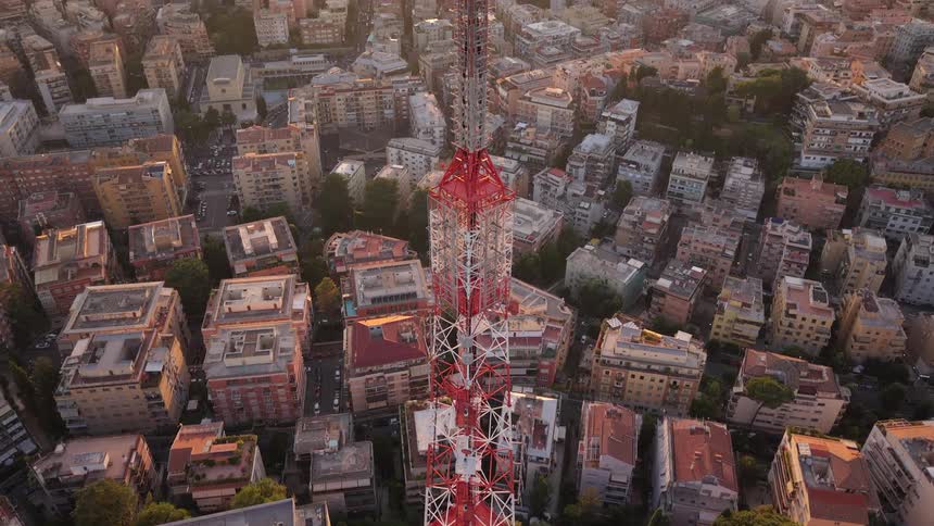 An upward tilting aerial view of a red and white radio tower reveals the expansive city skyline during a golden sunset