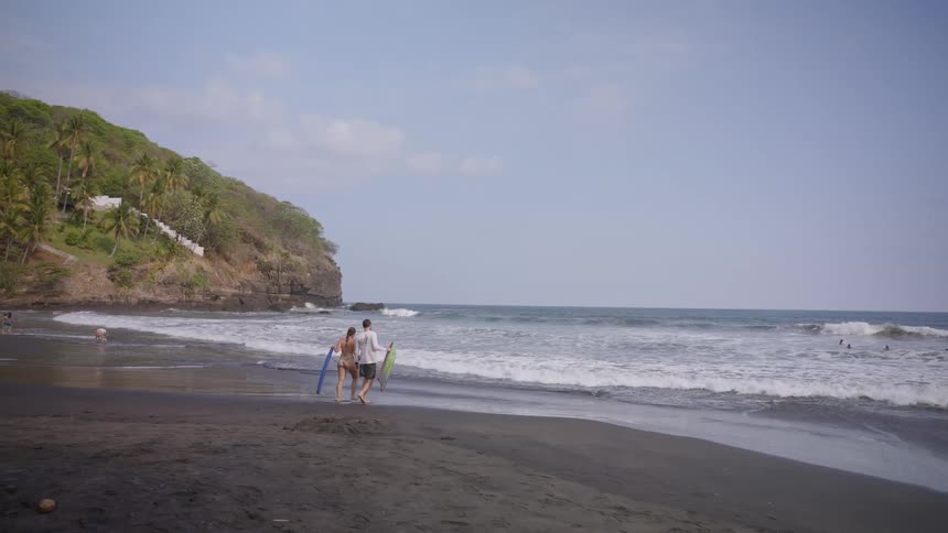 A couple with surfboards strolls along a scenic black sand beach as waves roll in