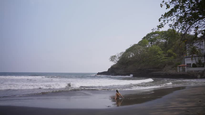 A woman sits in quiet contemplation on a dark volcanic sand beach as ocean waves wash ashore in slow motion