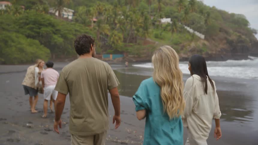 Rear view of a group of friends walking and talking on a dark sand beach next to the ocean on a cloudy day