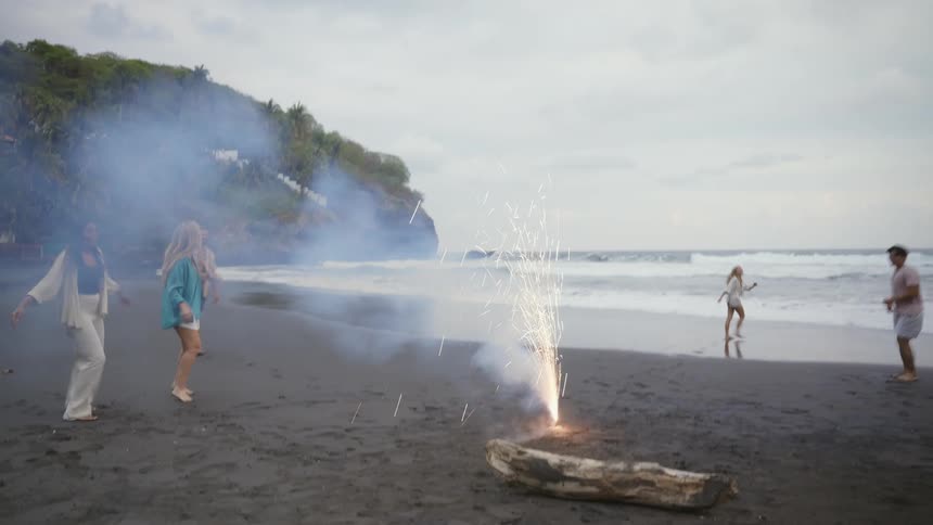 A group of diverse friends celebrates on a black sand beach, lighting a firework as one man playfully dances and does a backflip