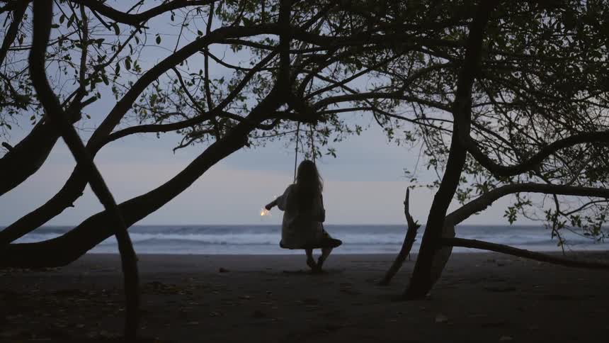 Silhouetted woman on a tree swing holds a lit sparkler and looks out at the ocean at dusk