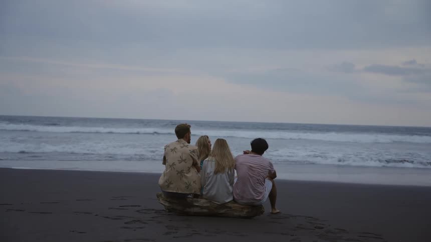 A group of four friends sits on a driftwood log, talking and enjoying the view of the ocean waves on a cloudy day