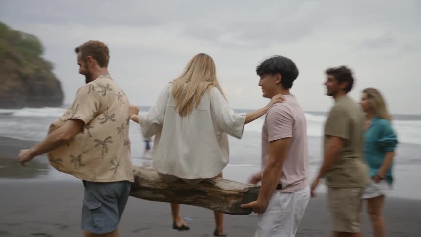 Two playful young men carry their friend on a large log as the group walks along the dark sand beach
