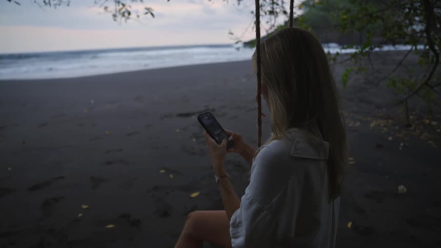 A woman relaxes on a rope swing at a dark sand beach in the evening while using her smartphone