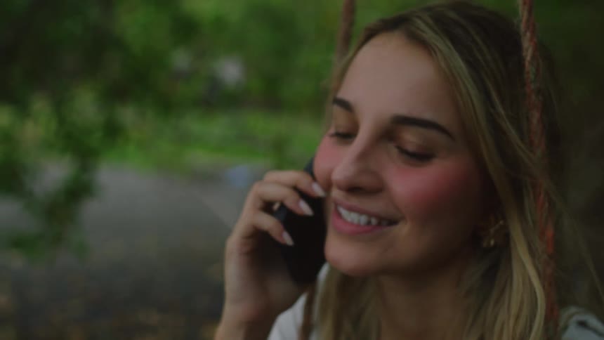 A joyful young woman with blonde hair smiles while talking on her phone on a swing outdoors
