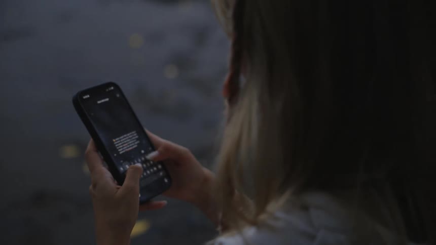 An over-the-shoulder close-up view shows a blonde woman's hands typing a message on her smartphone at dusk