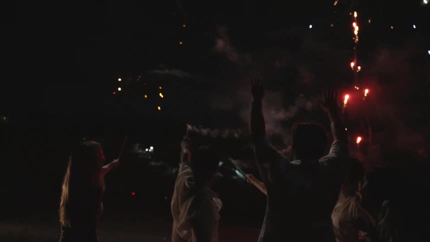 A diverse group of friends celebrate on the beach at night, setting off handheld fireworks and watching them explode in the sky