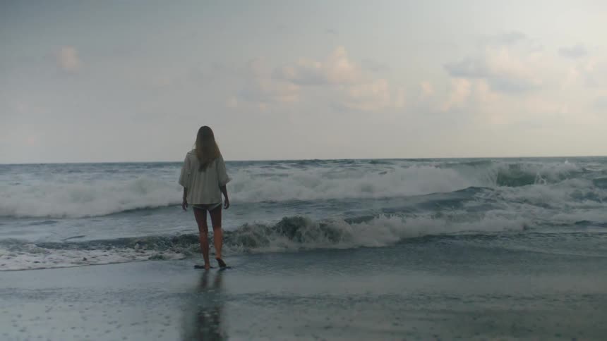 Slow motion of a contemplative woman standing barefoot on a sandy beach watching the waves crash