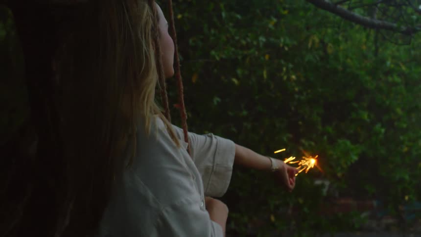 A serene young woman with long blonde hair enjoys a magical moment with a lit sparkler while sitting on a swing in a garden