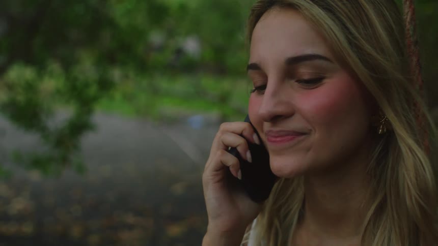 Close-up of a smiling young woman having a pleasant phone conversation while sitting on a swing outdoors