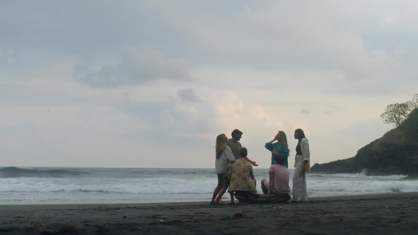 A group of young friends relax and socialize on a remote, dark sand beach as ocean waves crash on the shore under an overcast sky