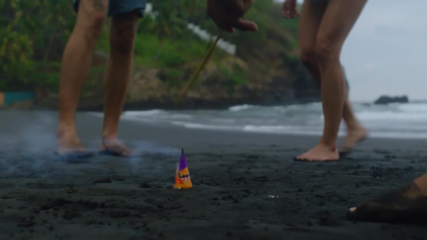 Close up slow motion shot of a hand lighting a fountain firework with a sparkler on a black sand beach as people run away