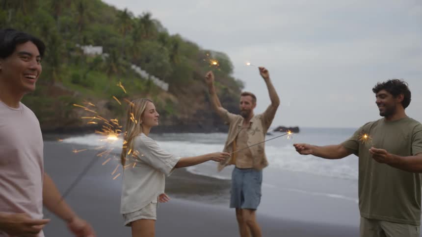 A group of diverse friends joyfully celebrate with lit sparklers on a dark sand beach, dancing and laughing in slow motion