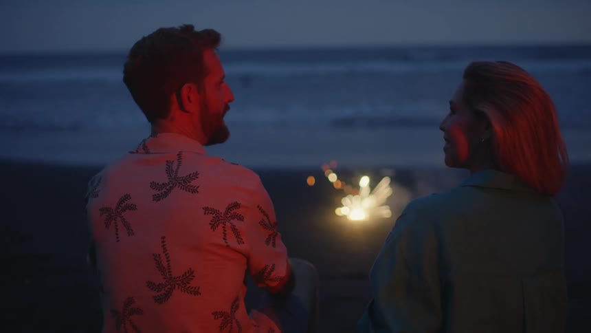 A romantic couple sits on the beach at night, talking and watching a sparkler on the sand in front of the ocean