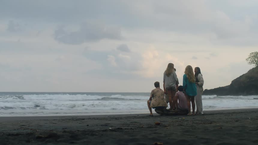 A group of young friends gather on a piece of driftwood, watching the waves roll in on a moody, overcast beach