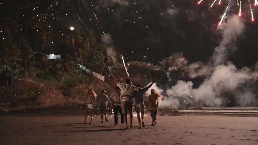 A group of friends celebrate on a tropical beach at night with a spectacular fireworks display